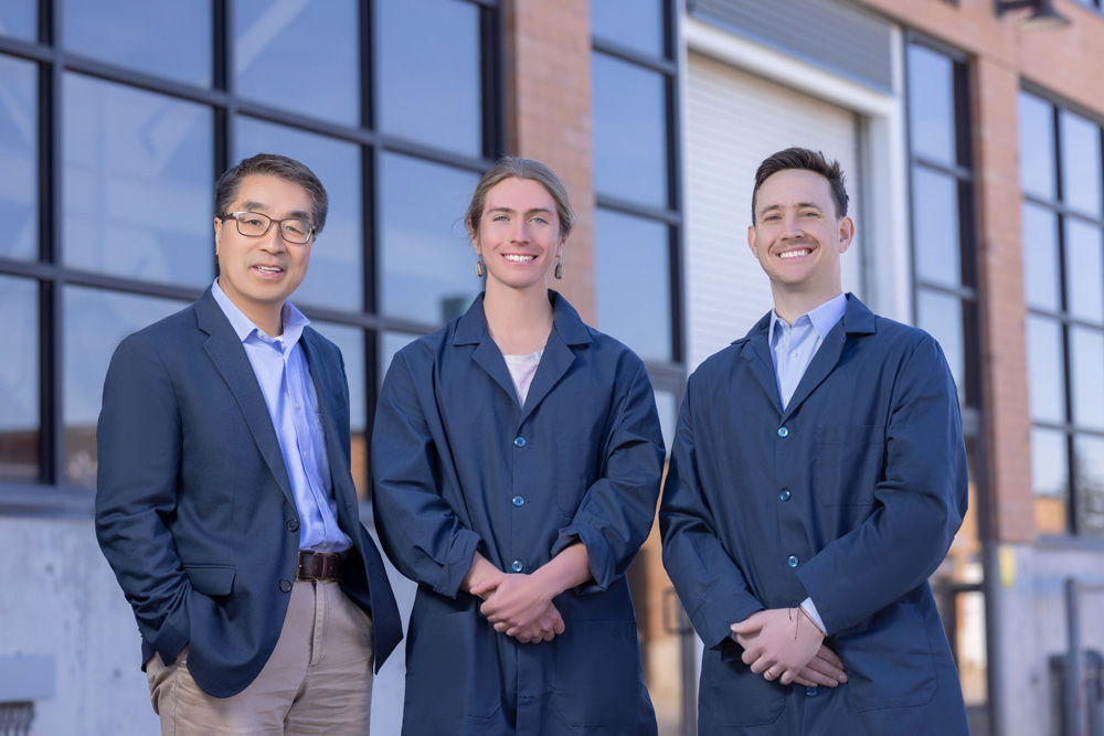 Dr. Zak Fang posing with two graduate student researchers in blue lab coats.