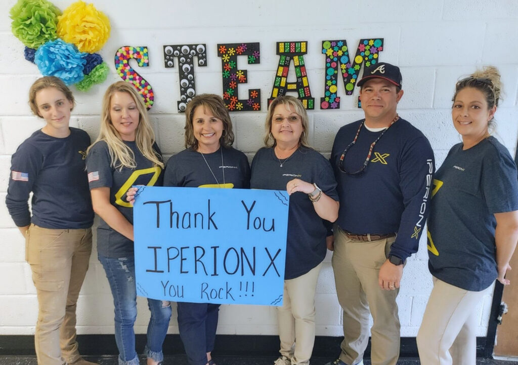 IperionX team members in west Tennessee holding a "Thank You IperionX You Rock!" sign for a STEAM classroom donation alongside educators in Camden, Tennessee.