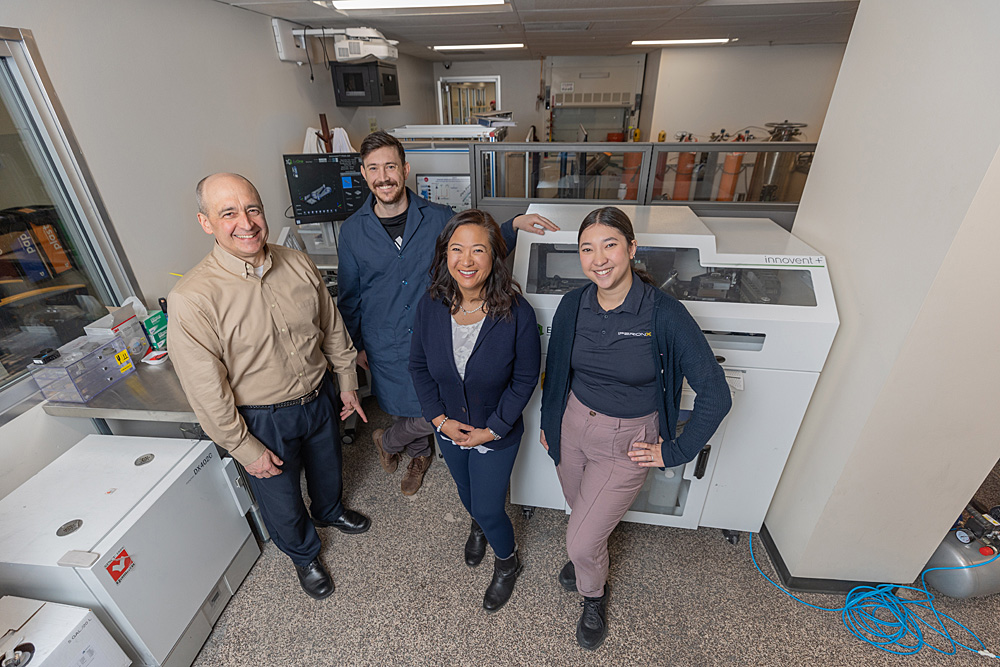 IperionX Head of Advanced Manufacturing, Harold Sears; graduate student in blue lab coat; IperionX Technical Advisor, Dr. Eliana Fu; and IperionX team member posing in front of binder jet 3D printer.